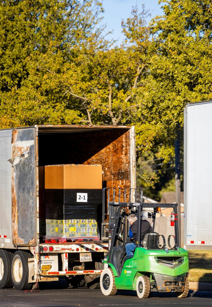 A forklift loading a truck with a pallet in an outdoor setting, surrounded by trees.