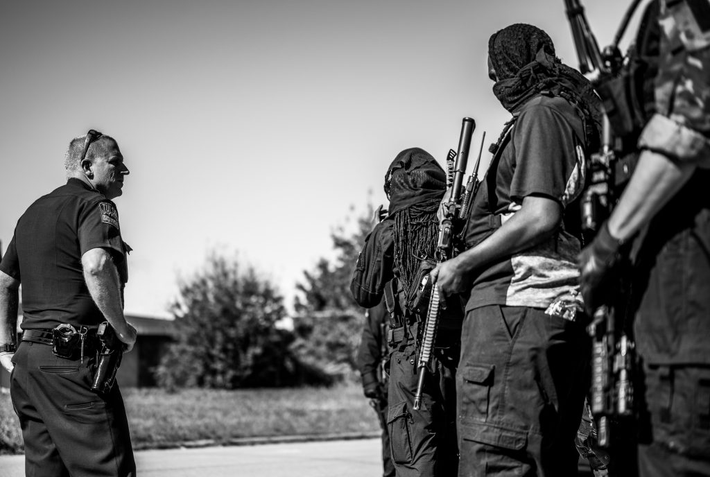 A group of armed individuals and police officer in black and white outdoor setting.