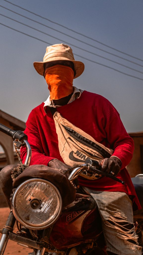 Person in red sweater and hat, face covered, sitting on a motorcycle outdoors.